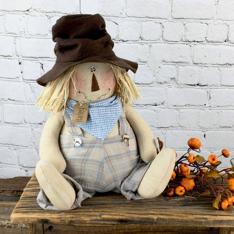 Rustic Harley the Scarecrow figurine wearing a hat and overalls, made from fabric and raffia, sitting on a farmhouse shelf with fall décor accents.