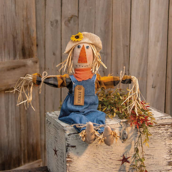 Patrick Scarecrow is a primitive doll that features raffia arms, legs, and hair for a realistic scarecrow finish. He wears a burlap hat with a yellow flower accent, an orange bandana, denim overalls, and a plaid shirt that is wrapped with jute at the wrists. He has a tag pinned to his chest that reads, "Patrick" and he looks great on a covered porch or seated around a hearth.