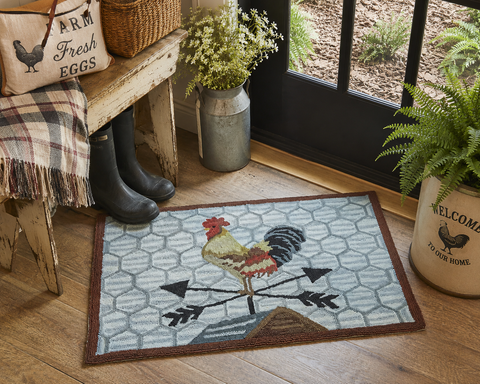 Break Of Day Rooster Hooked Rug by Teresa Kogut displayed beneath a wooden chair, featuring a colorful rooster standing on a weather vane against a chicken wire background. Rustic farmhouse hooked rug measuring 24 x 36 inches.