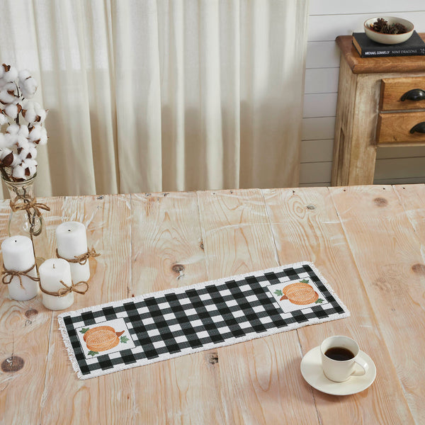 The Annie Black Check Pumpkin Runner measures 8 by 24 inches and features a black and white base in buffalo plaid surrounded by a soft, frayed edge. The left and right edges of the thanksgiving table runner feature a vintage-inspired pumpkin design in burnt orange with olive green leaves. 