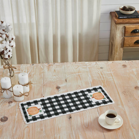 The Annie Black Check Pumpkin Runner measures 8 by 24 inches and features a black and white base in buffalo plaid surrounded by a soft, frayed edge. The left and right edges of the thanksgiving table runner feature a vintage-inspired pumpkin design in burnt orange with olive green leaves. 