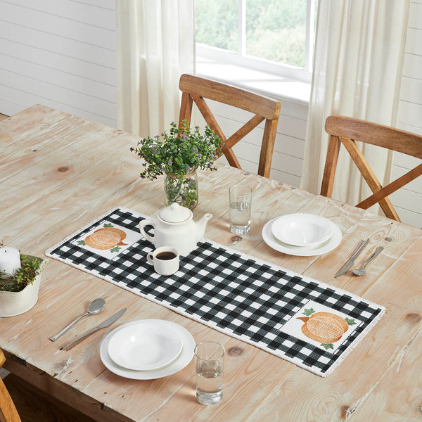 The Annie Black Check Pumpkin Runner measures 12 by 36 inches and features a black and white base in buffalo plaid surrounded by a soft, frayed edge. The left and right edges of the thanksgiving table runner feature a vintage-inspired pumpkin design in burnt orange with olive green leaves.