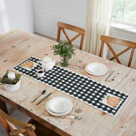 The Annie Black Check Pumpkin Runner measures 12 by 48 inches and features a black and white base in buffalo plaid surrounded by a soft, frayed edge. The left and right edges of the thanksgiving table runner feature a vintage-inspired pumpkin design in burnt orange with olive green leaves.