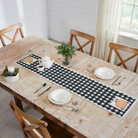 The Annie Black Check Pumpkin Runner measures 12 by 60 inches and features a black and white base in buffalo plaid surrounded by a soft, frayed edge. The left and right edges of the thanksgiving table runner feature a vintage-inspired pumpkin design in burnt orange with olive green leaves.