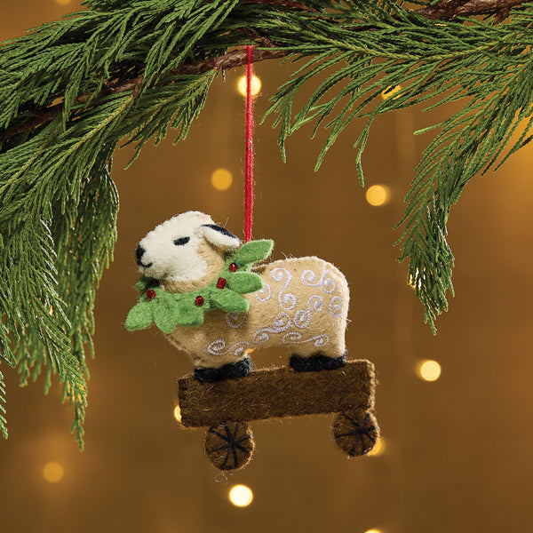 Sheep With Wreath Felt Ornament is a felt ornament depicting a sheep on wagon. The sheep is accented with white embroidery and a holly wreath with holly berries. Item is slightly filled for a weighted feel. Includes a string for hanging on the Christmas tree.