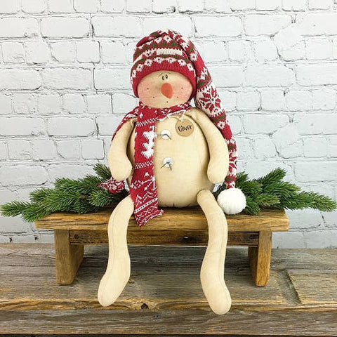 Oliver the Snowman figurine with long legs, wearing a red knit hat and scarf, sitting on a rustic wooden bench with holiday greenery.