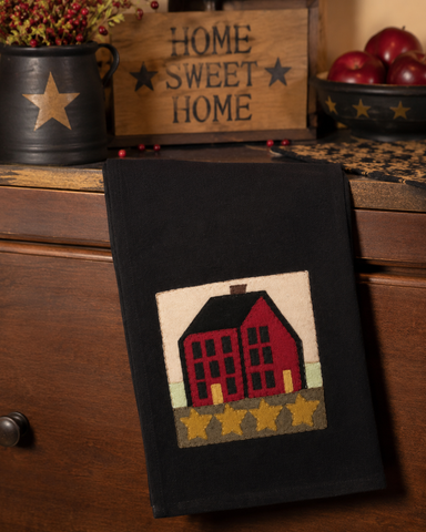 Black primitive kitchen towel featuring a red saltbox house appliqué with gold stars along the bottom border.