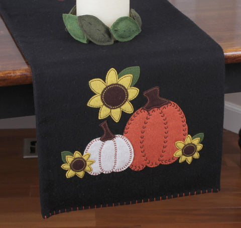 Black wool table runner with orange and white pumpkins, yellow sunflowers, and green leaves appliqué, accented with decorative stitching.