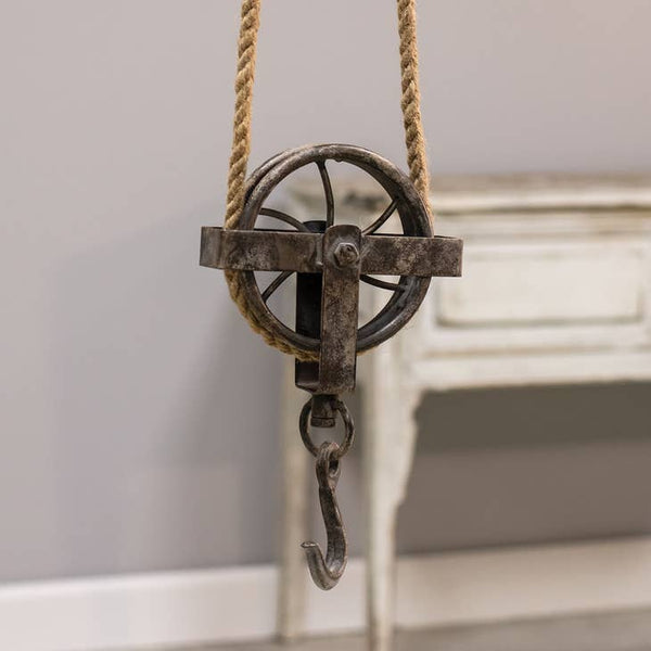 Pulley is made of worn-down metal with a primitive brown finish and hangs from a simple jute rope. The interior of the pulley is exposed so that wire spokes are visible.